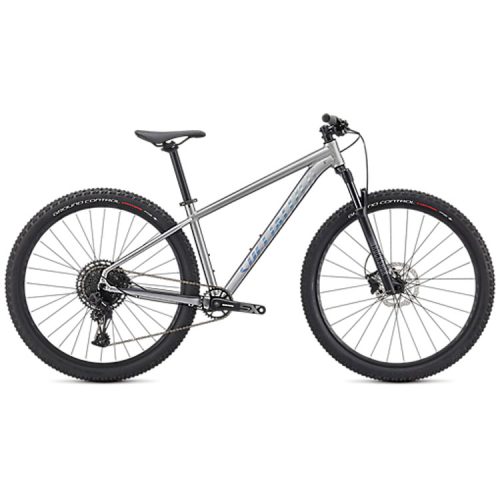 En silverfärgad mountainbike med svart styre, sadel och stora knobbiga däck, visad i profil mot en vit bakgrund. Cykeln har en framgaffel med fjädring och inga synliga tillbehör.