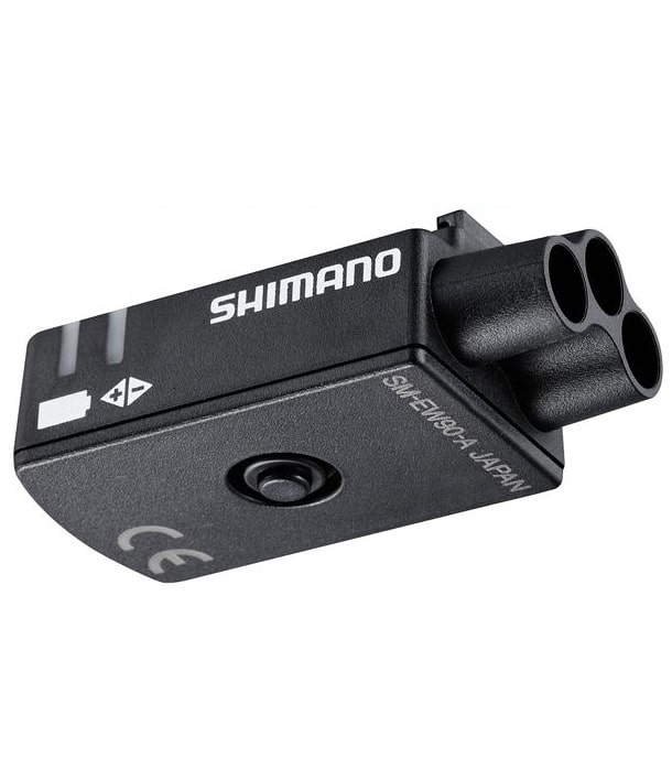 SM-EW90A.jpg En svart Shimano elektronisk cykelkomponent med tre cirkulära portar, en central knapp och vit textmärkning på sidan. Enheten har symboler och en CE-märkning på ytan.