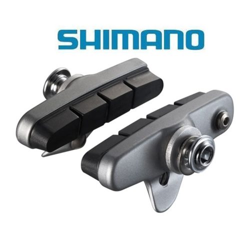 Två Shimano-bromsbelägg för cyklar visas mot en vit bakgrund med den blå SHIMANO-logotypen ovanför dem. Beläggen har en baksida av metall och bromsytor av svart gummi.