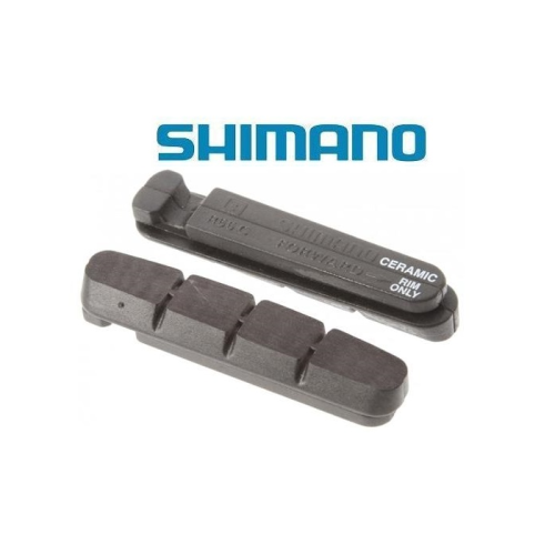 Två svarta Shimano-bromsbelägg för cyklar visas sida vid sida mot en vit bakgrund. Shimano-logotypen visas ovanför dem i blått. Det ena bromsbelägget är märkt "Ceramic Only".