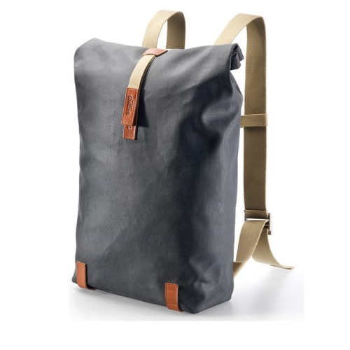 En minimalistisk mörkgrå ryggsäck med beige justerbara remmar, spänne på ovansidan och bruna läderdetaljer i övre och nedre hörnen, stående upprätt mot en vit bakgrund.
