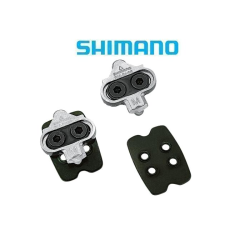 Ett par Shimano SPD-cykelklossar och två svarta rektangulära plattor visas på en vit bakgrund med den blå Shimano-logotypen ovanför dem.
