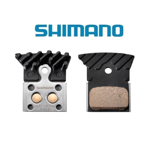 Två Shimano skivbromsbelägg för cykelbromsar visas sida vid sida. På det vänstra belägget syns metallstommen och kylflänsarna, medan friktionsmaterialet syns på det högra belägget. Shimanos logotyp visas ovanför beläggen.
