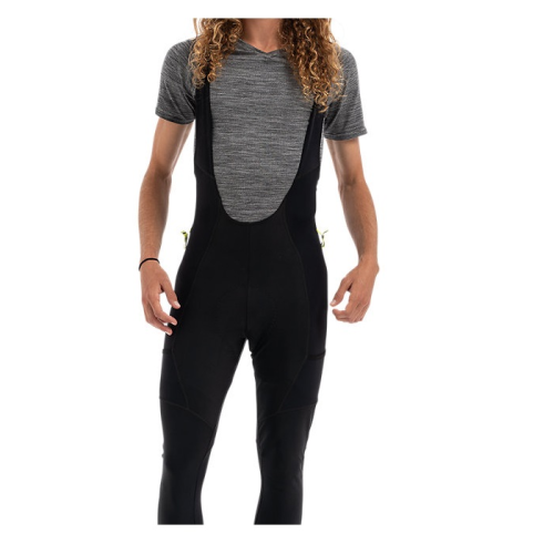 En person med långt lockigt hår bär en kortärmad grå skjorta och svarta cykeltights med haklapp. Huvudet är delvis utanför bild och bakgrunden är vit.