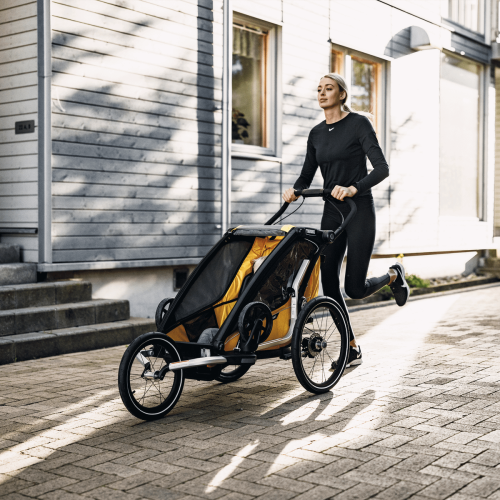 En blond kvinna i träningskläder joggar och skjuter en gul Thule Chariot barnvagn på en stengata. Soligt ljus skapar skuggor på husväggen i bakgrunden.