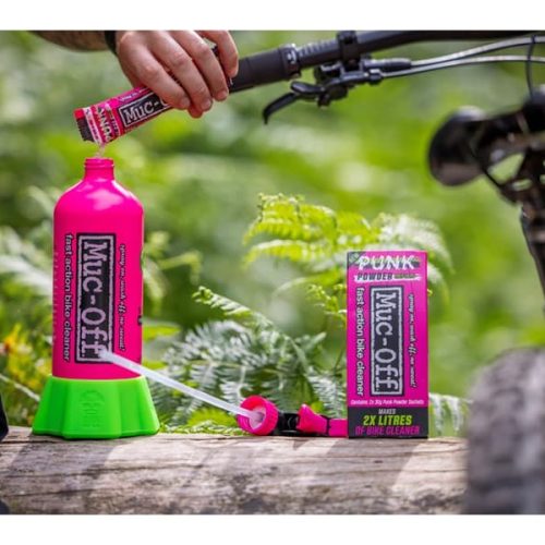 Utomhusbild visar en rosa flaska och förpackning av MUC-OFF Punk Powder Bike Cleaner. En hand häller pulvret i flaskan, medan en cykel står i bakgrunden. Scenen badar i naturligt ljus och förmedlar en aktiv och utomhusbetonad atmosfär.