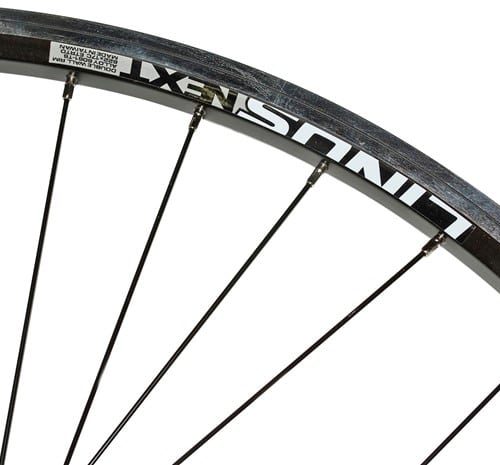 Detaljbild av ett svart cykelhjul med "LINUS NEXT" tryckt i vitt på fälgen. Ekrar löper mot en ljus bakgrund. Metalliska reflexer och skuggor antyder studioljus.