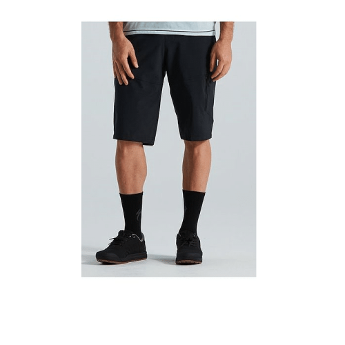 Endast den nedre halvan av en person är synlig, klädd i svarta Specialized Trail Cargo Shorts, en ljusgrå skjorta, svarta strumpor och svarta sportskor mot en vanlig ljus bakgrund.