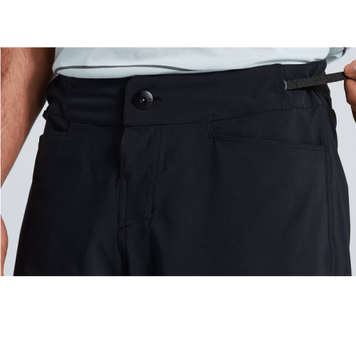 Närbild på en person som bär Specialized Trail Cargo Short; en hand drar i den justerbara midjebandsfliken och avslöjar shortsens främre knapp och fickor.