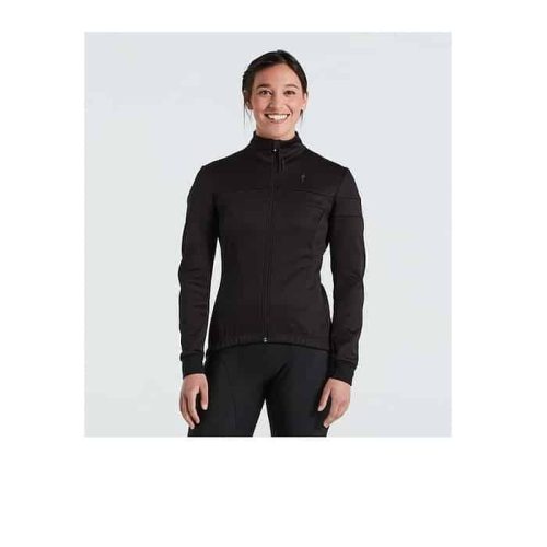 En kvinna står vänd mot kameran, ler och bär Specialized RBX Comp Softshell Jacket Women i svart mot en vanlig ljus bakgrund.