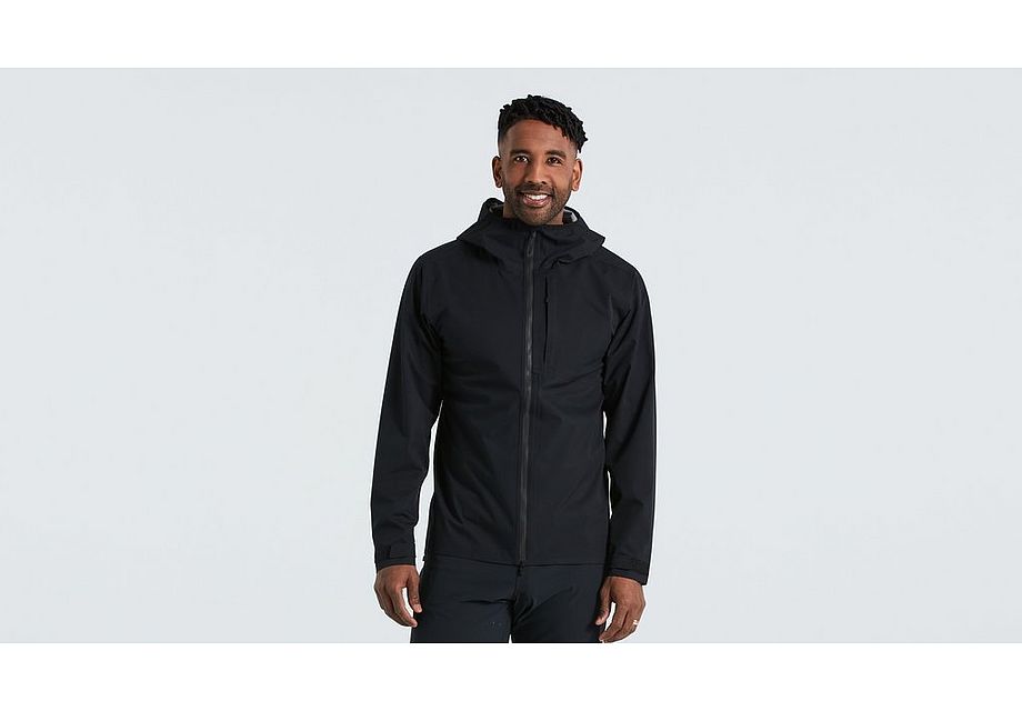 64422-810_.jpg En man klädd i Specialized Trail Rain Jacket i svart står vänd framåt mot en ljusgrå bakgrund och ler med händerna avslappnade i sidorna.