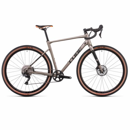 Gravel Cube Nuroad EX 1x11s - Brun är en silverfärgad gravelcykel med droppstyre, skivbromsar, breda däck med solbrända väggar och "CUBE"-märkning. Den har en sadel med orange accent och visas från sidan mot en vit bakgrund.