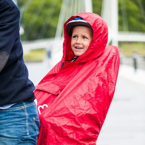 Ett barn i en röd Regnponcho Hamax och hjälm sitter leende bakom en vuxen på cykel, med suddiga träd och en bro i utomhusbakgrunden.
