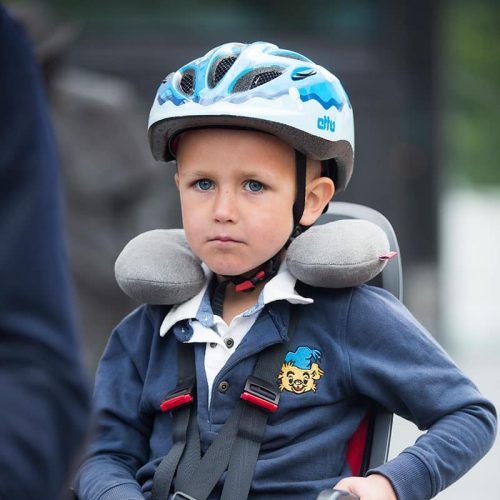 En ung pojke sitter i en barncykelsits, fastspänd med remmar, iklädd en blåvit hjälm och en marinblå jacka med en tecknad lapp. Han använder Nackkudde Hamax nackkudde och tittar rakt fram med ett allvarligt uttryck.