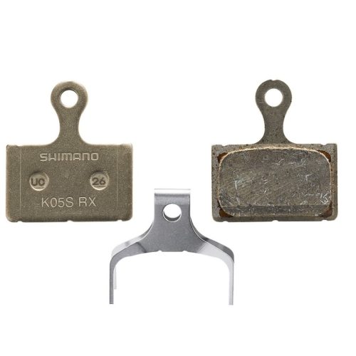 Närbild av Shimano K05S resin skivbromsbelägg, både nya och använda, samt en fjäder, mot en neutral bakgrund. Jämnt ljus framhäver detaljer som text och material.