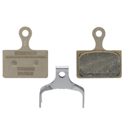 Närbild av Shimano K05Ti skivbromsbelägg för cyklar, resin-modell. Ett par belägg visas tillsammans med en fjäder. Detaljrikedom i texturer och material framhävs av jämn belysning.
