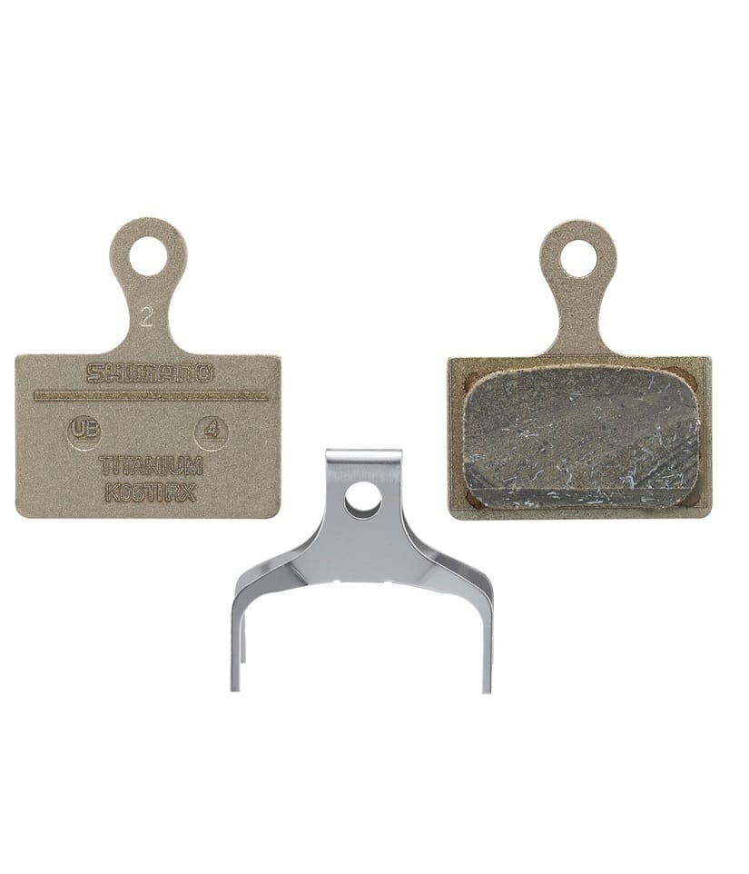 K05TI-RX-bel.jpg Närbild av Shimano K05Ti skivbromsbelägg för cyklar, resin-modell. Ett par belägg visas tillsammans med en fjäder. Detaljrikedom i texturer och material framhävs av jämn belysning.