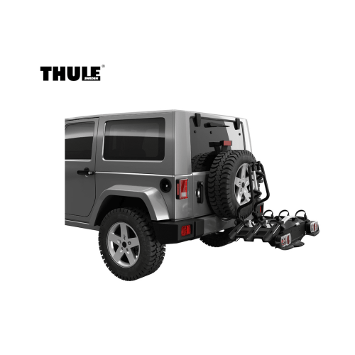 En silverfärgad SUV med ett bakmonterat reservhjul använder Adapter Thule VeloCompact Spare Wheel för att fästa ett Thule-cykelställ med två cyklar; Thule-logotypen visas i det övre vänstra hörnet på en vit bakgrund.