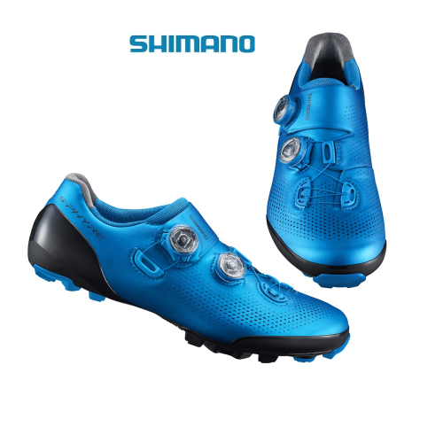 Ett par blå MTB-sko Shimano SH-XC901 S-Phyre cykelskor med tvåknäppning, perforerad ovandel, svart traktionssula och Shimano-logotypen ovanför, mot en vit bakgrund.