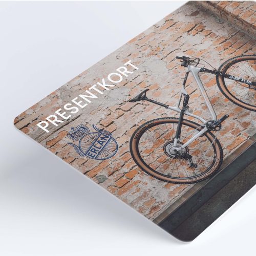 Detaljerad bild av ett Erlan Presentkort med en cykel framför en tegelvägg. Ljuset är jämnt fördelat, och fokus ligger på texten "Presentkort" och Erlan-logotypen. Atmosfären är modern och sportig.