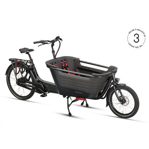 En svart Batavus Fier 2 Nexus lastcykel står i ett ljust och rent utrymme. Cykeln har en stor lastkorg fram med röda säkerhetsbälten och ett tydligt "3 års garanti" märke i övre högra hörnet.