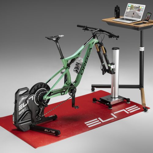 En detaljerad bild av en Climber Elite Rizer Backsimulator, komplett med cykel, laptop och tillbehör, placerad på en röd matta i en välbelyst miljö.