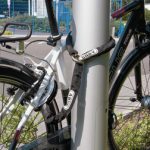 En ABUS ACH 6KS kätting låser fast två cyklar vid en silverfärgad stolpe i utomhusmiljö. Dämpad belysning framhäver låsets detaljer och cyklarnas konturer.