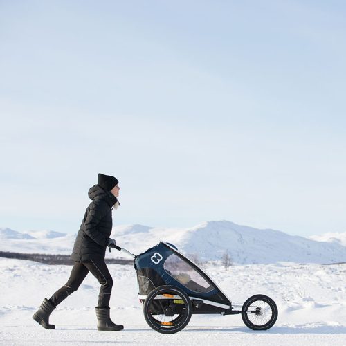 En kvinna i vinterkläder joggar med en Hamax Outback joggingkit i ett snöigt landskap med berg i bakgrunden. Bilden har ett starkt ljus och en kall atmosfär som betonar utomhusaktiviteten.