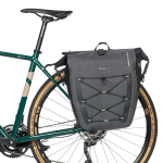 Närbild på en grön cykel med en grå Basil Navigator Storm sidoväska fäst på pakethållaren. Väskan har elastiska band och reflektiva detaljer, med bra ljus och fokus.