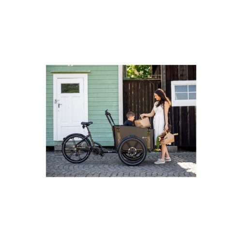 En kvinna står bredvid en Lastcykel Cargobike Delight, en trehjulig lastcykel med ett barn sittande i det främre utrymmet. Cykeln står parkerad på en kullerstensgata framför en grön byggnad med en vit dörr.