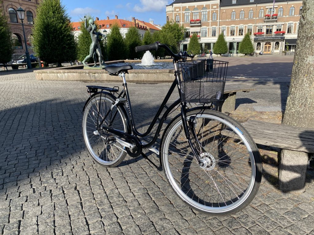 Svart Skeppshult Original cykel med korg, parkerad på kullersten intill en bänk. Fontän och byggnader i bakgrunden. Soligt väder med tydliga skuggor.