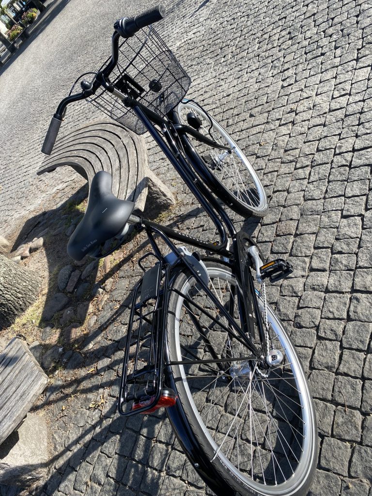 Svart Skeppshult Original cykel på kullersten, bredvid en bänk. Soligt väder och starka skuggor framhäver detaljerna på cykeln, inklusive korgen och sadeln.
