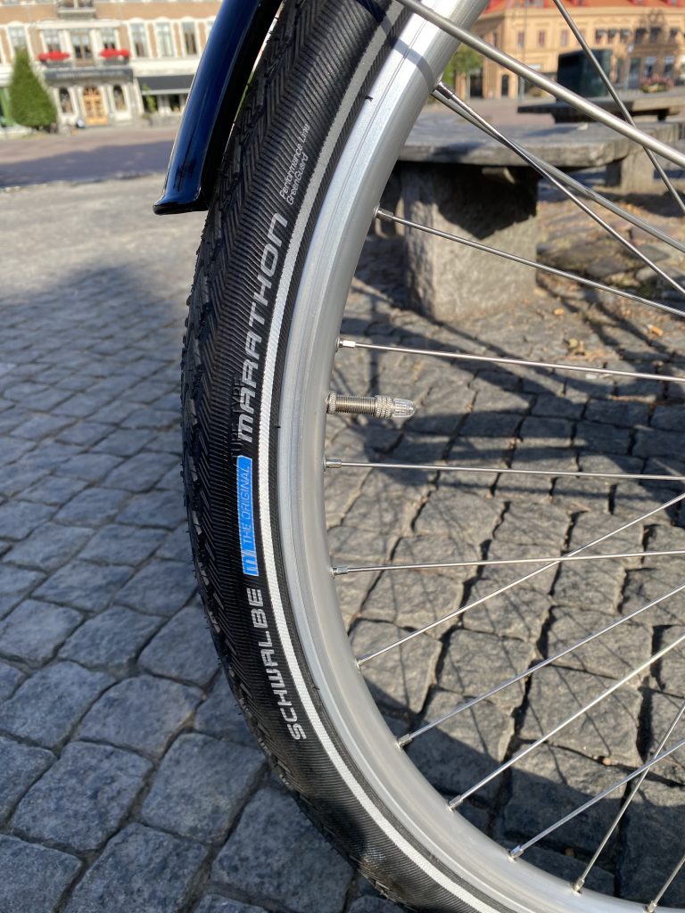 Närbild av ett cykelhjul med ett Schwalbe Marathon-däck mot en bakgrund av kullersten och en byggnad i fjärran. Ljuset är starkt och accentuerar detaljerna i däcket och hjulkonstruktionen.