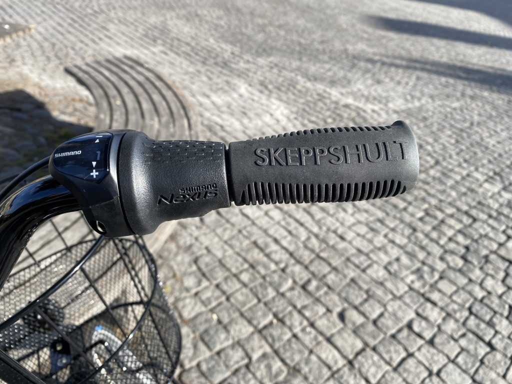 Närbild av ett svart cykelstyre med "SKEPPSHULT" präglat på handtaget och en Shimano Nexus-växel. Utebelysning framhäver strukturen på både handtaget och stenläggningen i bakgrunden.
