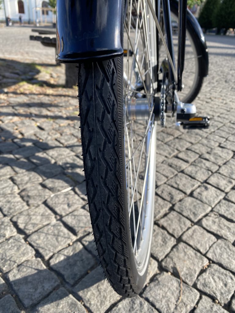 Detaljvy av ett Skeppshult Original cykeldäck på kullersten. Däcket är svart med ett distinkt mönster, och metallfälgen glänser i det soliga ljuset. Bilden framhäver cykelns robusta konstruktion.