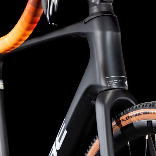 Närbild av en Cube Cross Race C:62 PRO cykel, med fokus på framgaffeln och däcket Schwalbe X-One. Det mattsvarta chassit framhävs av stark belysning mot en svart bakgrund.
