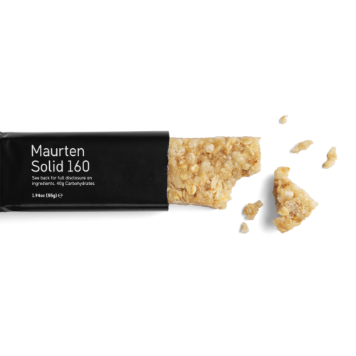 Maurten Solid 160 energibar, delvis uppäten, ligger på en vit bakgrund. Den svarta förpackningen kontrasterar mot den ljusa energibaren, som har en smulig konsistens. Mindre bitar av baren ligger spridda runt omkring.