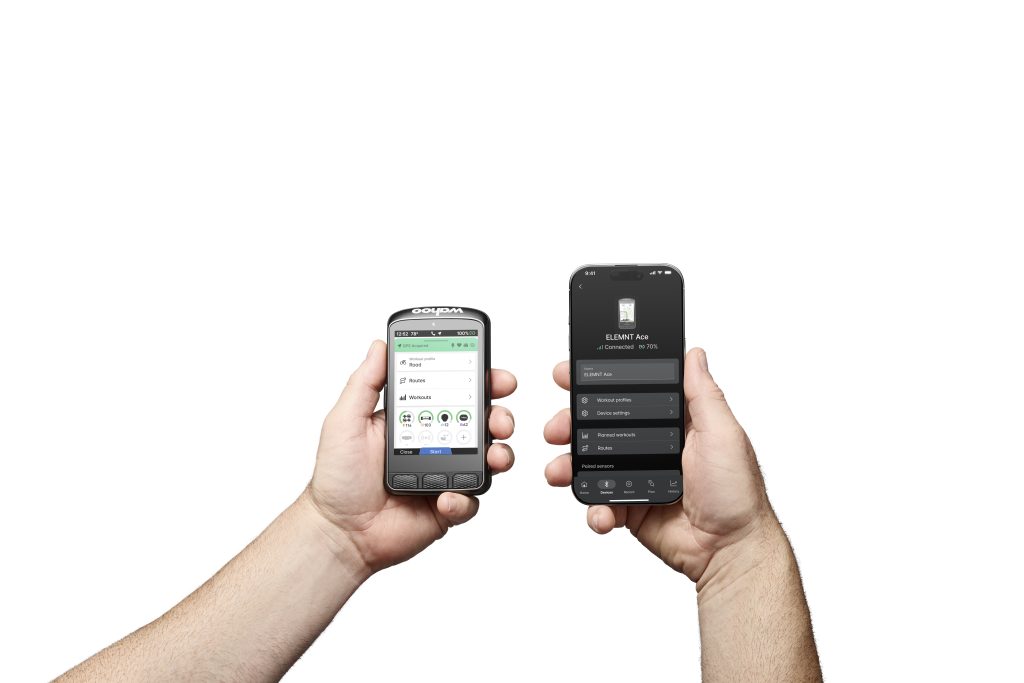 Två händer håller en Wahoo ELEMNT Ace och en iPhone som visar en app som synkroniserar information mellan enheterna. Ljuset är starkt, vilket framhäver detaljerna i enheterna och händerna.