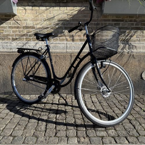 Svart Damcykel Skeppshult Original 3s med korg framför tegelvägg och kullersten.