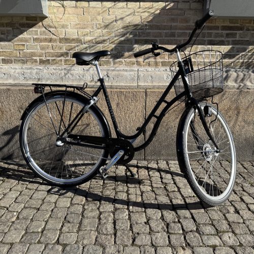 Svart Skeppshult Original 3s Damcykel med korg, parkerad mot en vägg.