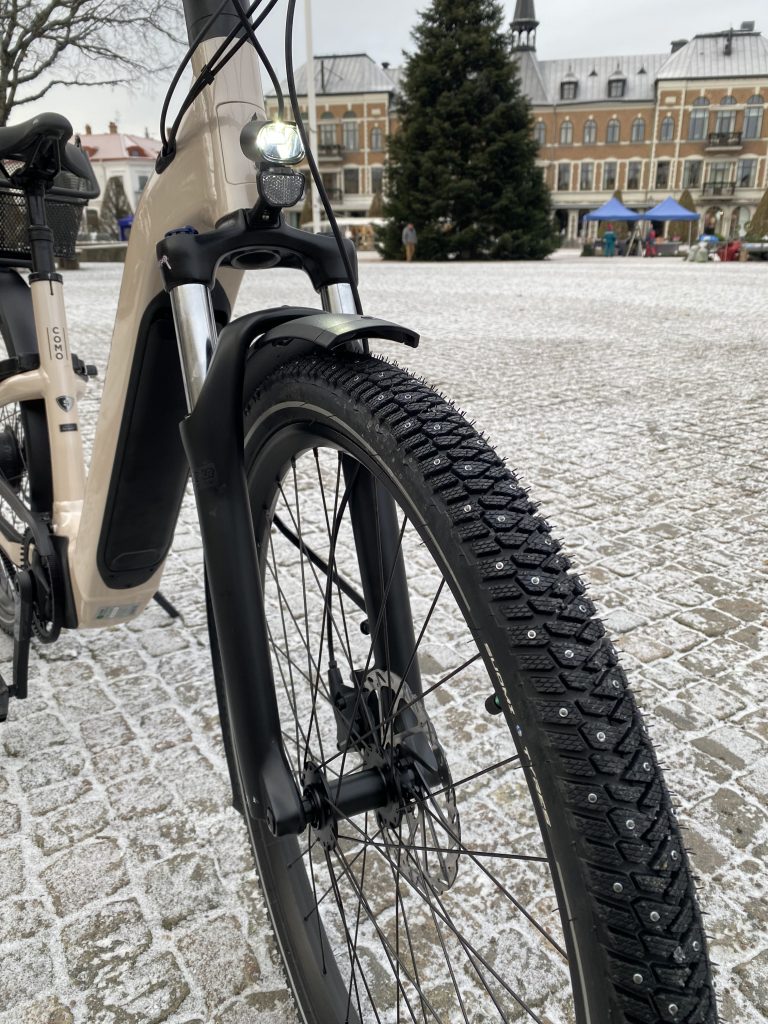 Närbild av framhjulet på en beige cykel med dubbdäck, parkerad på en snöig kullerstensyta. En julgran och historiska byggnader bildar bakgrunden-Guide: hitta de bästa vinterdäcken för din cykel.