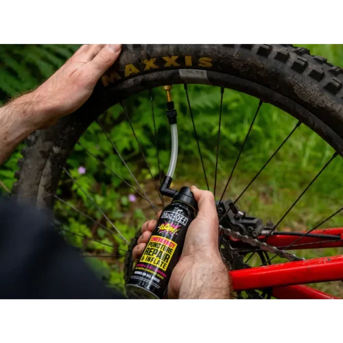Någon använder Punkteringslagning MUC-OFF B.A.M. Bottled Air Magic - 125 ml för att reparera och pumpa upp ett MAXXIS mountainbikedäck på en röd cykel, med gröna bladverk i bakgrunden.
