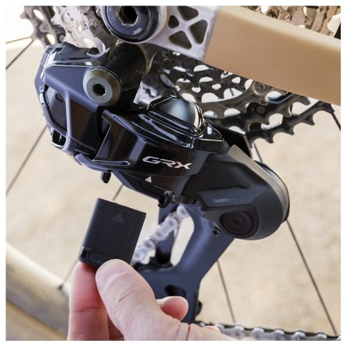 En hand sätter in ett litet rektangulärt batteri i bakväxeln Shimano GRX RD-RX827 SGS Di2 1x12s derailleur monterad på en cykels bakhjul, med växlar och kedja synliga i bakgrunden.