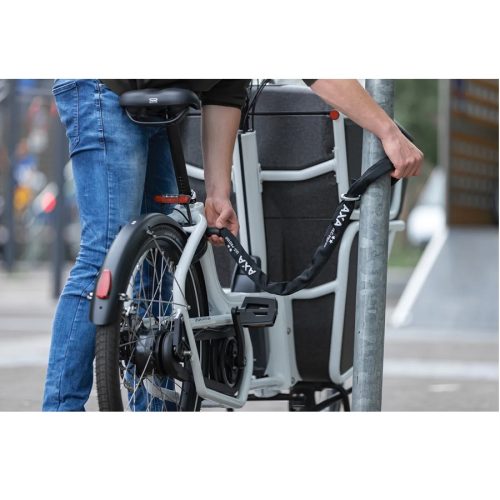 En person i blåjeans fäster en cykel på en metallstolpe med hjälp av en Kätting AXA ULC Pro Plug-in låskätting 8 mm kätting x 100 cm.
