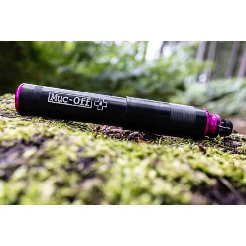 En MUC-OFF AirMach minipump (7,6 bar/110 psi) i svart och rosa vilar på grön mossa utomhus, med suddiga träd och bladverk i bakgrunden.
