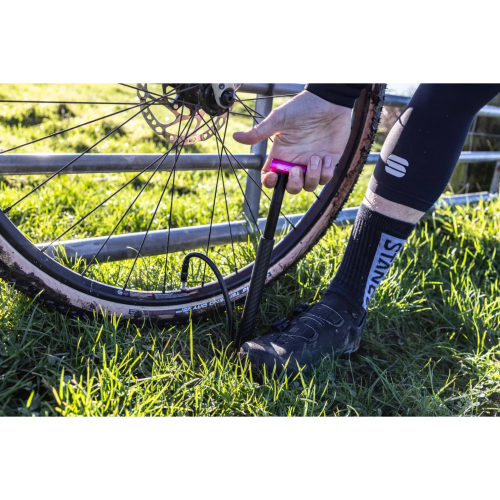 Iförd svarta cykelskor, strumpor och tights använder en person en MUC-OFF Minipump Airmach Carbon 130 psi Svart/rosa för att pumpa upp det leriga framdäcket på en cykel på gräs.