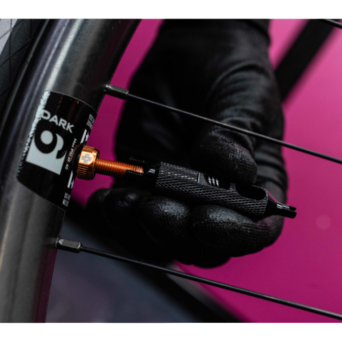 En hand i svart handske använder Ventilverktyg MUC-OFF - Valve Core Remover på ett cykelhjuls svarta och orange ventilskaft nära en "DARK"-etikett. Bakgrunden är rosa.