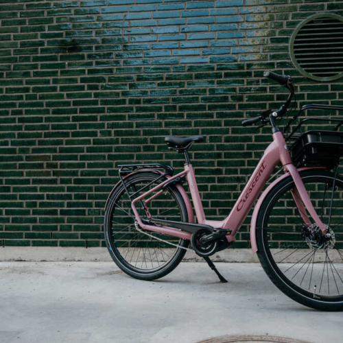 En rosa cykel med en korg fram och ett pakethållare bak står parkerad på en betongyta framför en grön kaklad vägg med en rund ventilationsöppning.