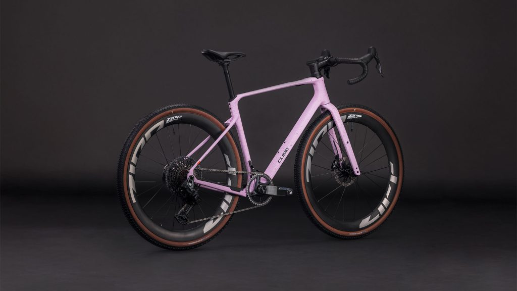 Gravel CUBE Nuroad C:62 SLX, 1x12s - Rosa är en ljusrosa gravelcykel med droppstyre, tjocka terrängdäck, skivbromsar, lättviktsram och fälgar med djupa profiler mot en mörk bakgrund.