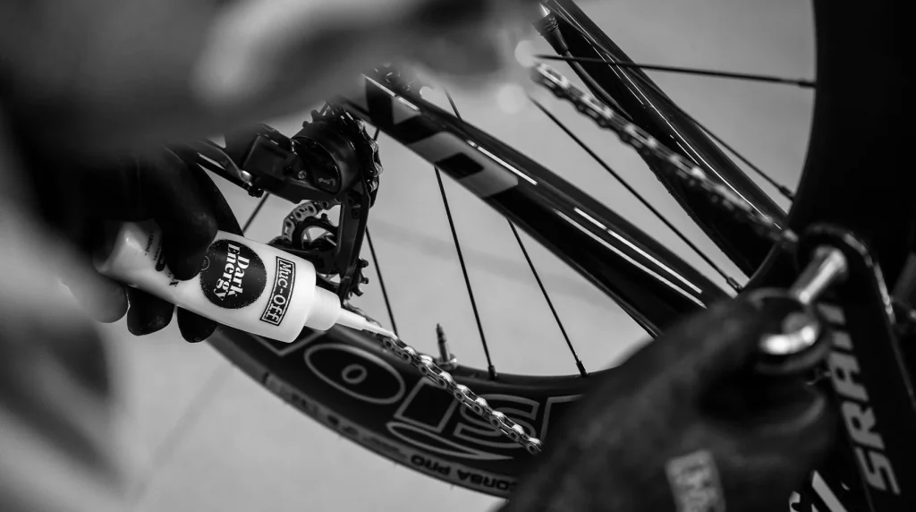 En person med handskar smörjer kedjan på en cykel, med fokus på drivlinan och bakhjulet. Bilden är i svartvitt.